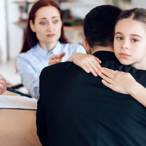 Sad daughter hugs dad who is sitting at the table by the lawyer. Young parents came to the office of a lawyer to file a divorce and share property.